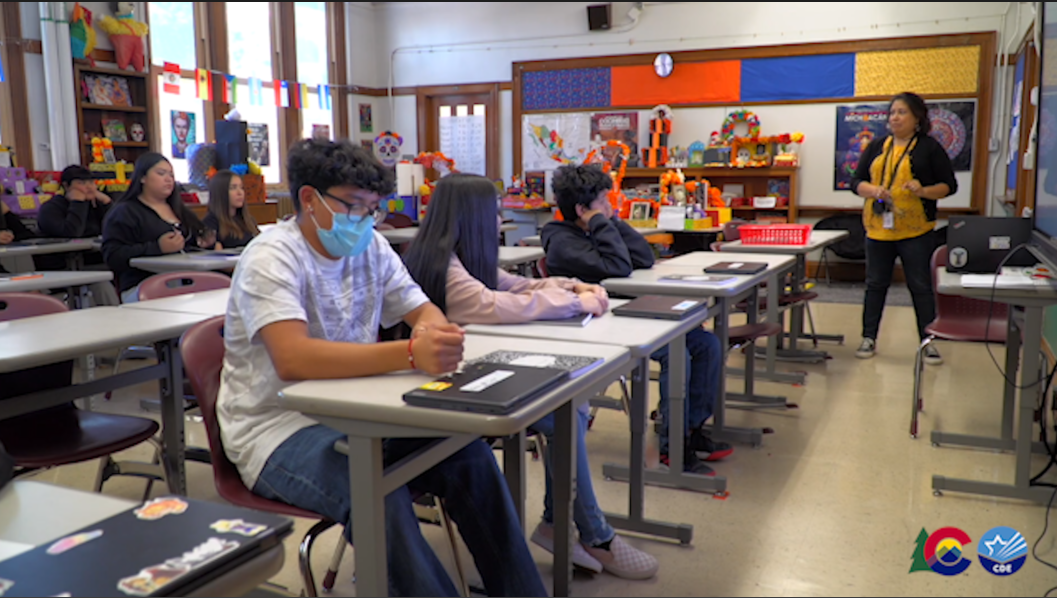 Students sit at desks as a teacher stands in the front of a classroom. 