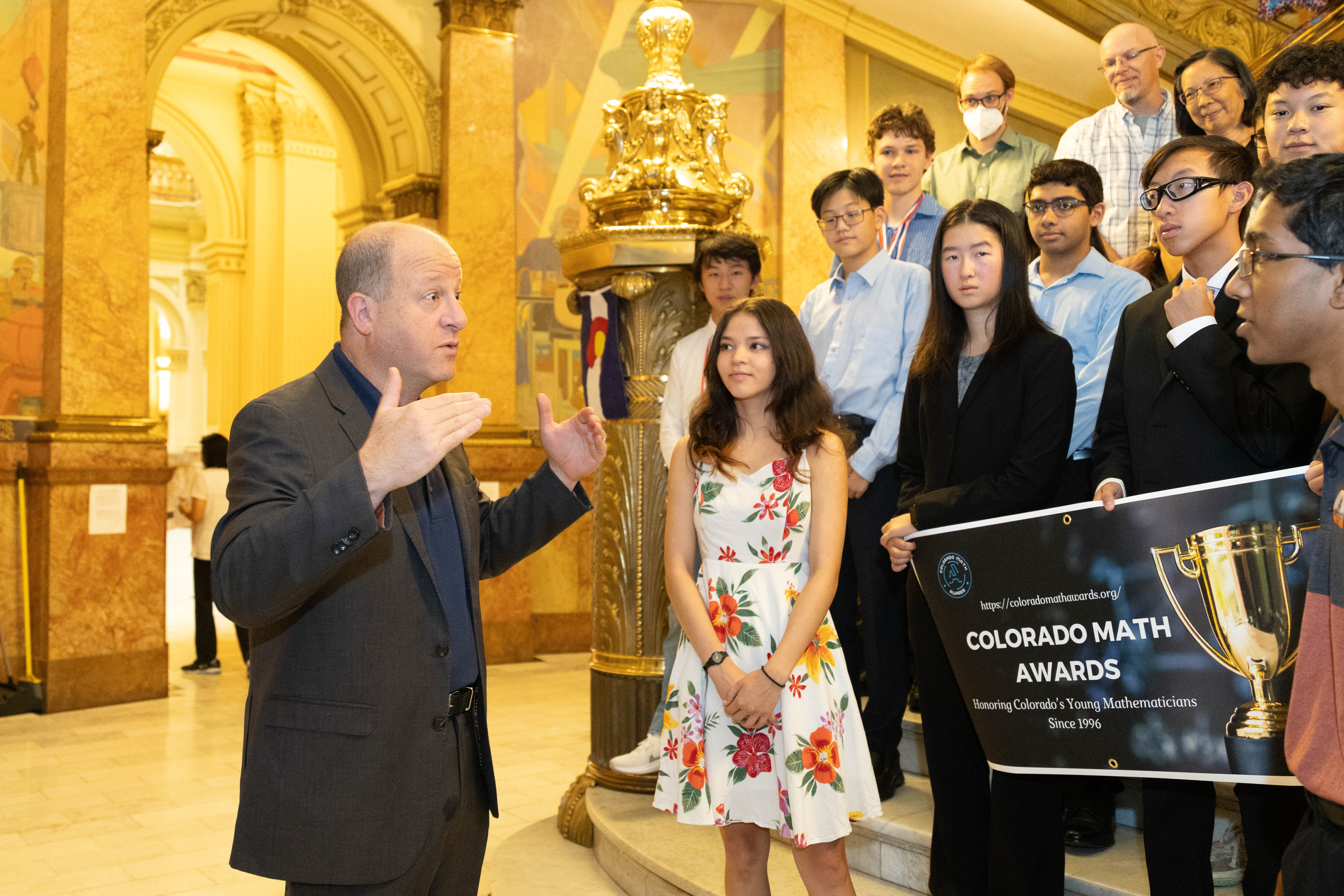 Gov. Jared Polis speaks to a group of students holding a Colorado Math Awards banner.