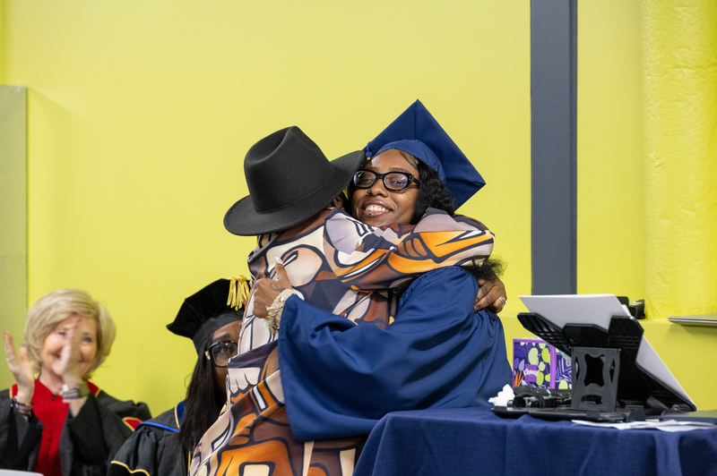  A woman in a blue cap and gown smiles and hugs another woman.