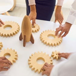 Hands placing wooden gears on a table around a small wooden rocketship.