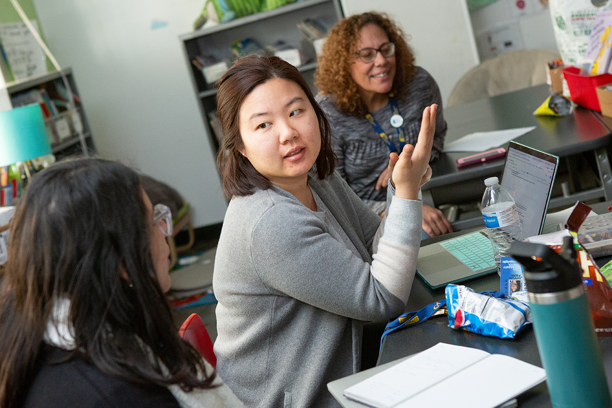 Group of adults in a classroom setting having a discussion, focus is on a woman who is speaking while another woman smiles in the background